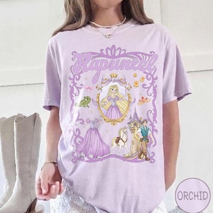 May include: A lilac t-shirt with a Rapunzel graphic. The design includes Rapunzel in a gold frame, a frog, a tower, and other characters. The word "Rapunzel" is written in a decorative font.