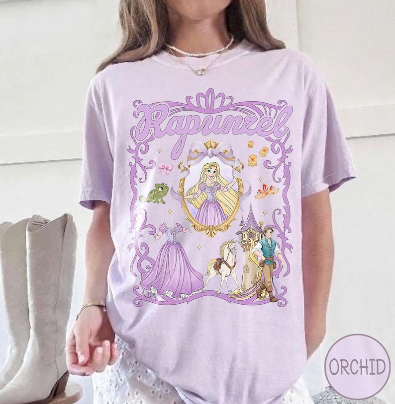 May include: A lilac t-shirt with a Rapunzel graphic. The design includes Rapunzel in a gold frame, a frog, a tower, and other characters. The word "Rapunzel" is written in a decorative font.