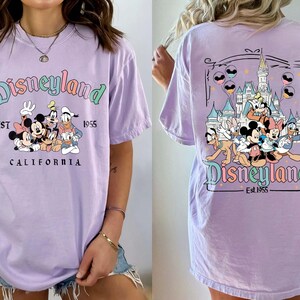 Może przedstawiać: Liliowa koszulka z logo Disneyland i grafiką Myszki Miki, Myszki Minnie, Kaczora Donalda i Goofy'ego. Prz&oacute;d koszulki przedstawia logo Disneyland i napis "EST 1955 CALIFORNIA". Tył koszulki ma grafikę zamku.
