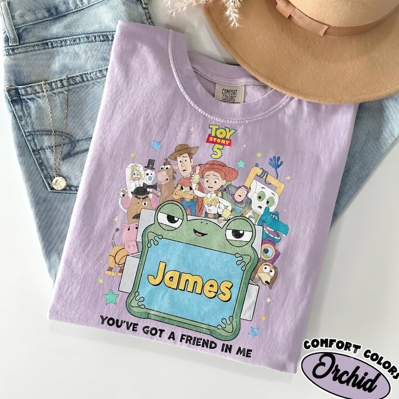 May include: A light purple t-shirt featuring a Toy Story 5 graphic with various characters, a large green frog illustration, and the name James printed in yellow. The text You've Got A Friend In Me is printed below the frog. The shirt has a crew neckline.