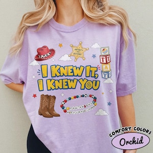 Pode incluir: Uma t-shirt cor de orqu&iacute;dea com uma impress&atilde;o gr&aacute;fica com o texto "I KNEW IT, I KNEW YOU". O design inclui um chap&eacute;u de cowboy vermelho, uma estrela de xerife dourada, blocos de letras, botas de cowboy castanhas e uma pulseira de contas com o texto "You've got a friend in me".