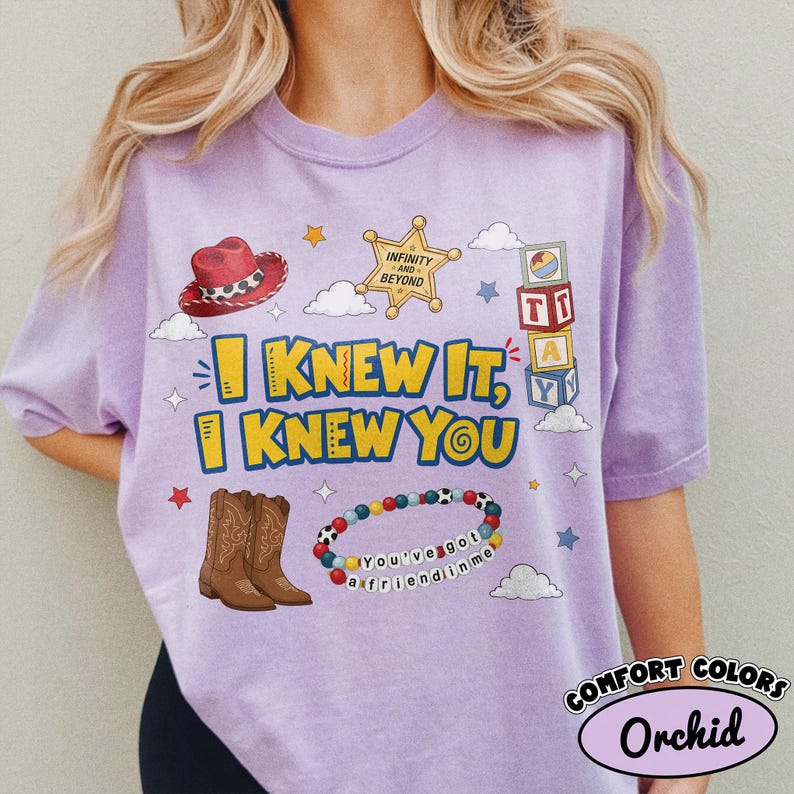 Pode incluir: Uma t-shirt cor de orqu&iacute;dea com uma impress&atilde;o gr&aacute;fica com o texto "I KNEW IT, I KNEW YOU". O design inclui um chap&eacute;u de cowboy vermelho, uma estrela de xerife dourada, blocos de letras, botas de cowboy castanhas e uma pulseira de contas com o texto "You've got a friend in me".
