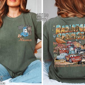 Puede incluir: Camiseta verde musgo con un dise&ntilde;o de coche de dibujos animados y el texto "Name" en la parte delantera. La parte trasera de la camiseta muestra "Radiator Springs" con varios personajes de coches y la frase "Welcome to Radiator Springs".