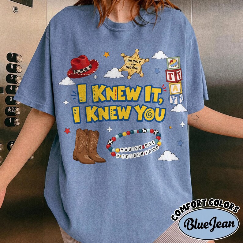 Pode incluir: Uma t-shirt azul com uma estampa gr&aacute;fica com o texto I KNEW IT, I KNEW YOU. O design inclui um chap&eacute;u de cowboy vermelho, uma estrela de xerife amarela, blocos de letras empilhados, botas de cowboy castanhas e uma pulseira de contas com o texto YOU'VE GOT A FRIEND IN ME.