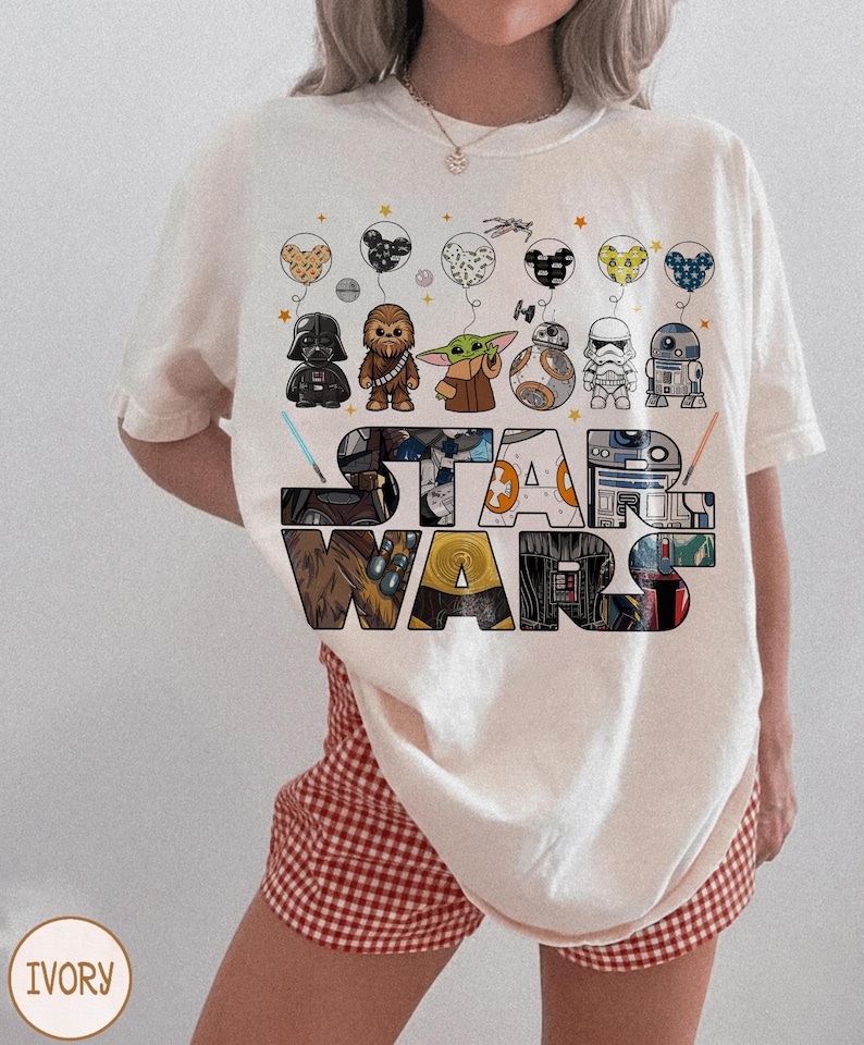 May include: Ivory t-shirt with the words "STAR WARS" in a colourful, block letter design. Above the text are illustrated balloons with Star Wars characters. The shirt has a relaxed fit.