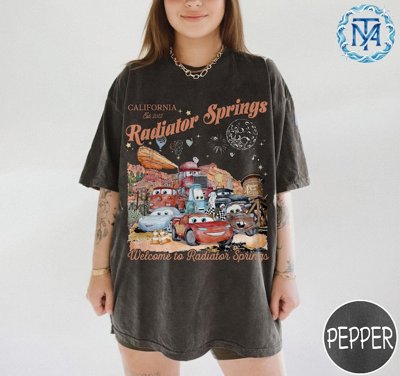 May include: Dark gray t-shirt featuring a graphic of the town of Radiator Springs from the movie Cars. The design includes the text "California Est. 2012 Radiator Springs" and "Welcome to Radiator Springs". The graphic features several cartoon cars.