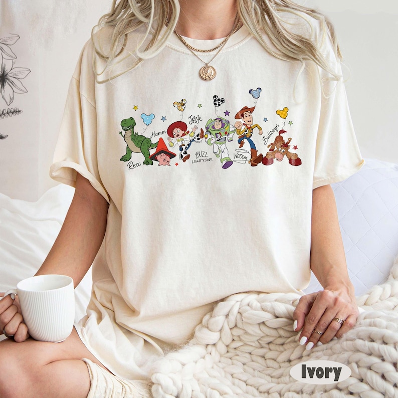 May include: Ivory-coloured t-shirt with a colourful graphic of Toy Story characters, including Rex, Hamm, Jessie, Buzz Lightyear, Woody, and Bullseye. The names of the characters are printed below them. The shirt is a relaxed fit.