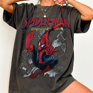 May include: Dark grey t-shirt with a Spider-Man graphic. The design features the text "SPIDER-MAN" in red and "BRAND NEW DAY". The superhero is shown in his red and blue suit, striking a pose against a cityscape background.
