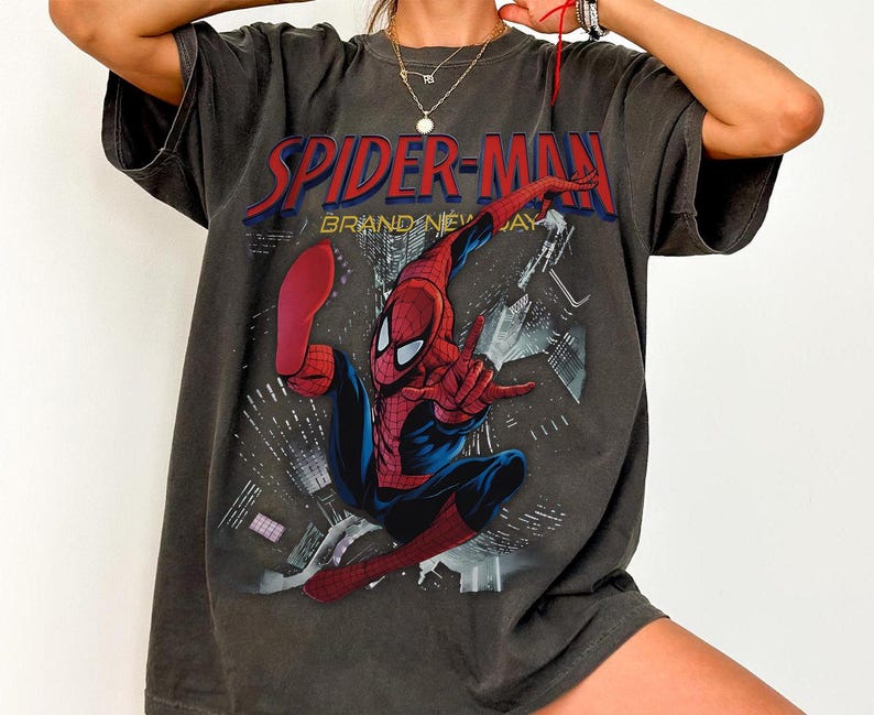 May include: Dark grey t-shirt with a Spider-Man graphic. The design features the text "SPIDER-MAN" in red and "BRAND NEW DAY". The superhero is shown in his red and blue suit, striking a pose against a cityscape background.