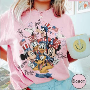 May include: Pale pink t-shirt with a patriotic Disney design featuring Mickey, Minnie, Donald, Daisy, Goofy, and Pluto, with American flags and fireworks. A mug with a smiley face is held in the image. The text includes "USA 1776-2026".