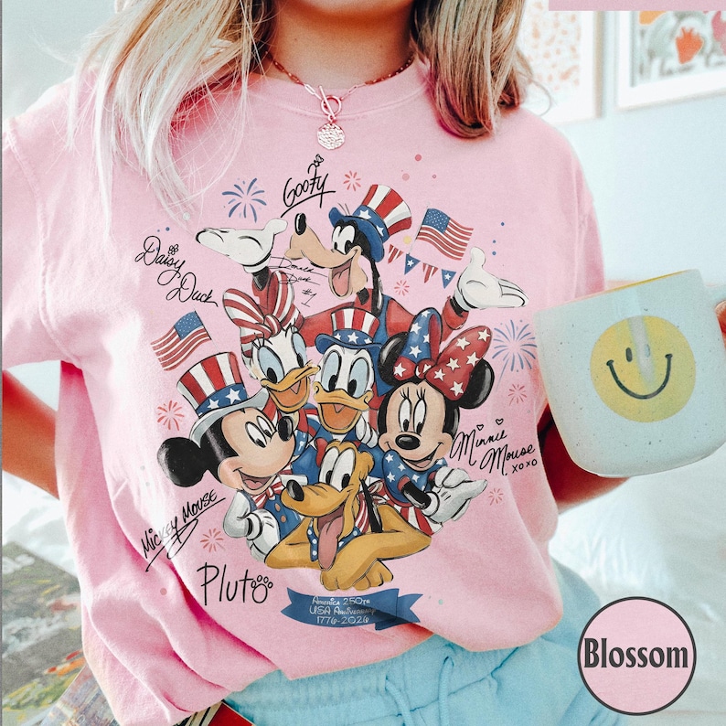 May include: Pale pink t-shirt with a patriotic Disney design featuring Mickey, Minnie, Donald, Daisy, Goofy, and Pluto, with American flags and fireworks. A mug with a smiley face is held in the image. The text includes "USA 1776-2026".