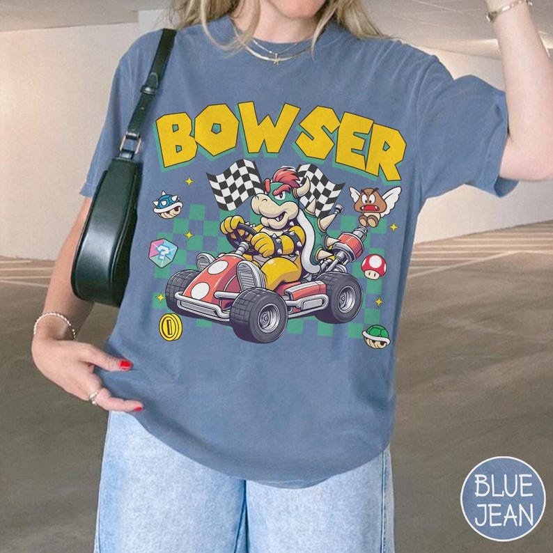 May include: A blue t-shirt featuring a graphic of Bowser driving a red go-kart with checkered flags. The design includes yellow text reading BOWSER, a green checkered background, a gold coin, a mushroom, a turtle shell, and a winged Goomba.
