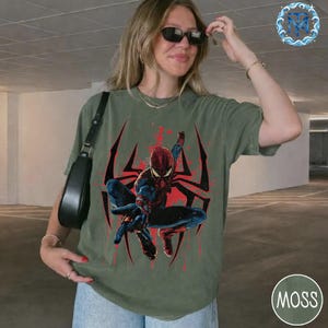 Puede incluir: Camiseta verde oliva con un dise&ntilde;o de Spider-Man en una pose din&aacute;mica, sobre un fondo de telara&ntilde;a roja y negra. El superh&eacute;roe lleva su traje cl&aacute;sico rojo y azul. La camiseta tiene un corte casual y holgado.