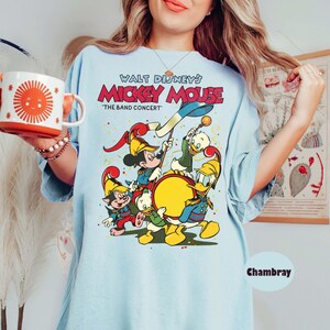 May include: Light blue t-shirt featuring a colorful graphic of Mickey Mouse and friends in a band, with the text "Walt Disney's Mickey Mouse 'The Band Concert'". The shirt has a relaxed fit and short sleeves.