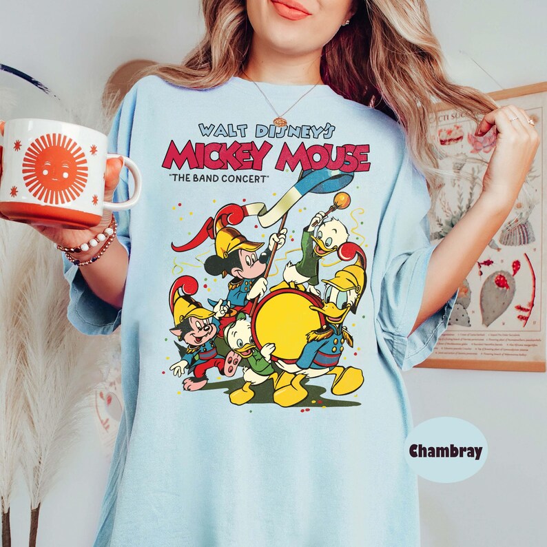 May include: Light blue t-shirt featuring a colorful graphic of Mickey Mouse and friends in a band, with the text "Walt Disney's Mickey Mouse 'The Band Concert'". The shirt has a relaxed fit and short sleeves.