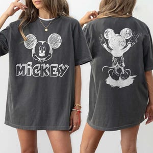 May include: Dark grey t-shirt with a white Mickey Mouse design on the front and back. The front displays Mickey's face and the word "Mickey". The back shows Mickey with his arms raised in a peace sign. A casual, unisex garment.