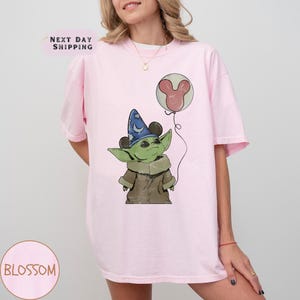 Puede incluir: Camiseta rosa claro con una ilustraci&oacute;n de dibujos animados de un personaje alien&iacute;gena verde que lleva un sombrero de mago y sostiene un globo de Mickey Mouse. La camiseta tiene el texto "Blossom" en un c&iacute;rculo en la parte inferior.