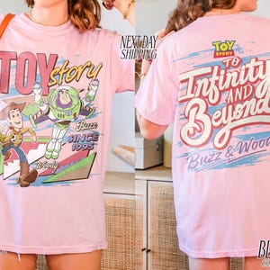 K&ouml;nnte beinhalten: Hellrosa T-Shirt mit Toy Story-Grafik, die Woody und Buzz Lightyear zeigt. Auf der R&uuml;ckseite steht der Schriftzug "To Infinity and Beyond". Das T-Shirt ist aus Comfort Colors Material.