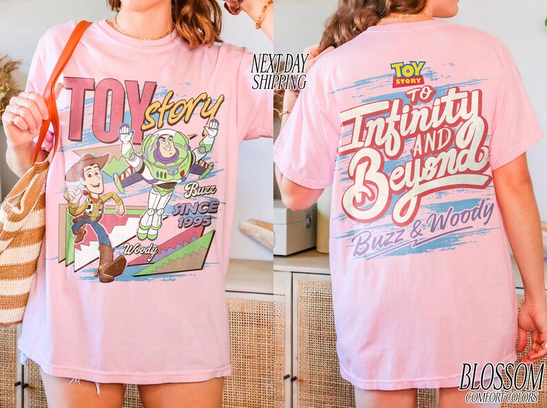 K&ouml;nnte beinhalten: Hellrosa T-Shirt mit Toy Story-Grafik, die Woody und Buzz Lightyear zeigt. Auf der R&uuml;ckseite steht der Schriftzug "To Infinity and Beyond". Das T-Shirt ist aus Comfort Colors Material.