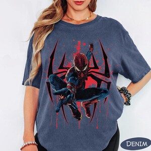 Puede incluir: Camiseta azul denim con un gr&aacute;fico de Spider-Man en una pose din&aacute;mica, sobre un dise&ntilde;o de telara&ntilde;a roja y negra. El superh&eacute;roe est&aacute; representado con su cl&aacute;sico traje rojo y azul. La palabra "DENIM" est&aacute; impresa en la parte inferior.