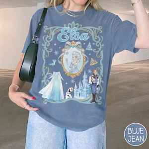 May include: Blue jean-coloured t-shirt featuring a graphic of Elsa from Frozen. The design includes Elsa's name, a portrait, dress, castle, and characters. The shirt has a round neck and short sleeves.