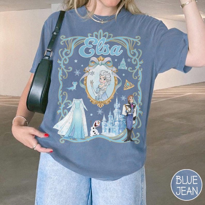 May include: Blue jean-coloured t-shirt featuring a graphic of Elsa from Frozen. The design includes Elsa's name, a portrait, dress, castle, and characters. The shirt has a round neck and short sleeves.