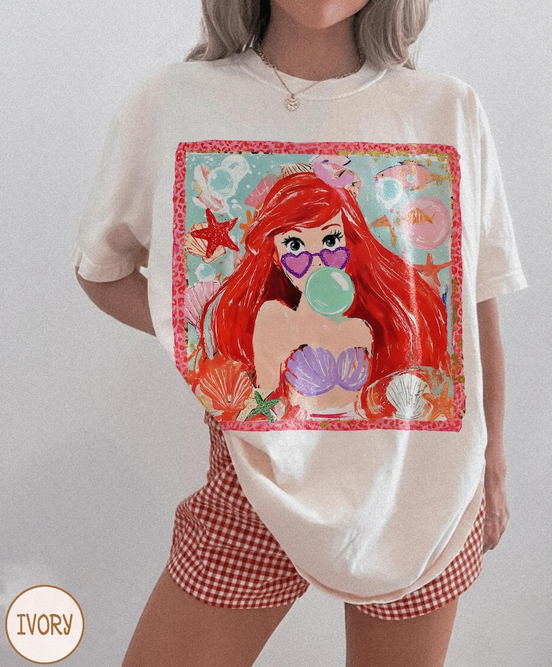 Puede incluir: Camiseta de color marfil con un gr&aacute;fico colorido de una sirena con pelo rojo, gafas de sol en forma de coraz&oacute;n y una burbuja. El dise&ntilde;o incluye conchas marinas, estrellas de mar y peces. La camiseta se combina con pantalones cortos a cuadros rojos y blancos.