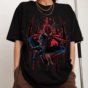 Puede incluir: Camiseta negra con un gr&aacute;fico de Spider-Man en una pose din&aacute;mica. El superh&eacute;roe est&aacute; representado con su cl&aacute;sico traje rojo y azul, sobre un dise&ntilde;o de telara&ntilde;a roja. El dise&ntilde;o incluye efectos de salpicaduras de pintura roja.