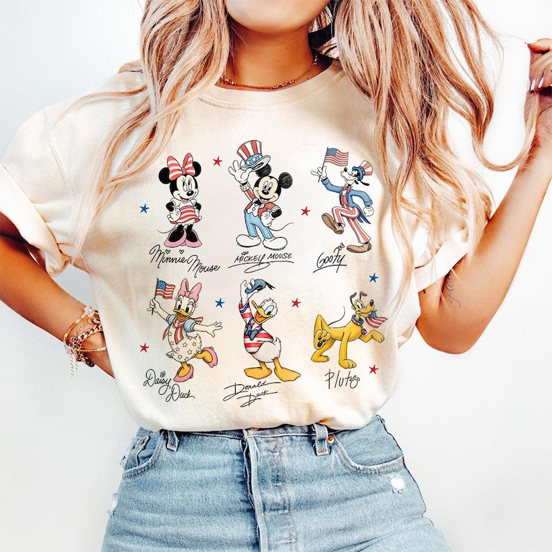 May include: Cream-coloured t-shirt featuring cartoon characters Minnie Mouse, Mickey Mouse, Goofy, Daisy Duck, Donald Duck, and Pluto. Each character is depicted in patriotic attire, holding flags or wearing festive hats. The characters' names are written below them.