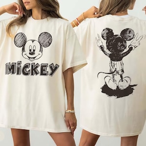May include: Ivory-coloured t-shirt with a black and white Mickey Mouse graphic. The front shows Mickey's face with the name "Mickey" in a 3D font. The back features a full-body sketch of Mickey Mouse with his arms raised in a peace sign.