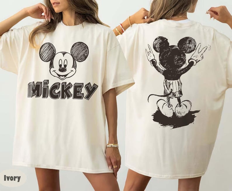 May include: Ivory-coloured t-shirt with a black and white Mickey Mouse graphic. The front shows Mickey's face with the name "Mickey" in a 3D font. The back features a full-body sketch of Mickey Mouse with his arms raised in a peace sign.
