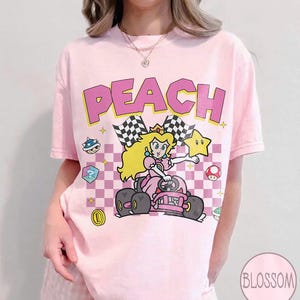 May include: A light pink t-shirt featuring a graphic print of Princess Peach driving a go-kart. The design includes the text PEACH in large pink letters, a checkered background, a yellow star, a blue shell, a mushroom, and a gold coin.
