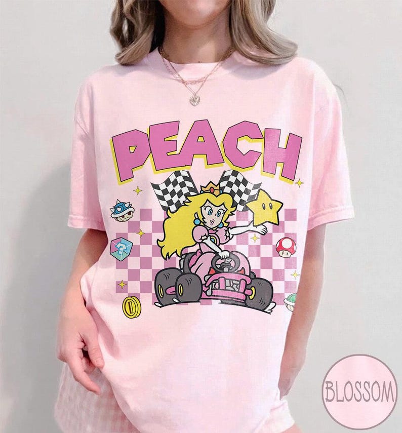 May include: A light pink t-shirt featuring a graphic print of Princess Peach driving a go-kart. The design includes the text PEACH in large pink letters, a checkered background, a yellow star, a blue shell, a mushroom, and a gold coin.