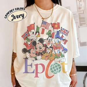 Puede incluir: Una camiseta de color marfil con un gráfico de Mickey Mouse, Minnie Mouse, Goofy, el Pato Donald, la Pata Daisy y Pluto rodeados de varias banderas internacionales. El texto Epcot aparece impreso bajo los personajes con un globo terráqueo en la o.