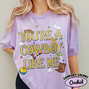Puede incluir: Una camiseta de manga corta de color morado claro con el texto amarillo "YOU'RE A COWBOY LIKE ME" rodeado de gr&aacute;ficos que incluyen un sombrero de vaquero marr&oacute;n, una insignia de sheriff, un cohete, un alien&iacute;gena verde, una pelota y nubes.