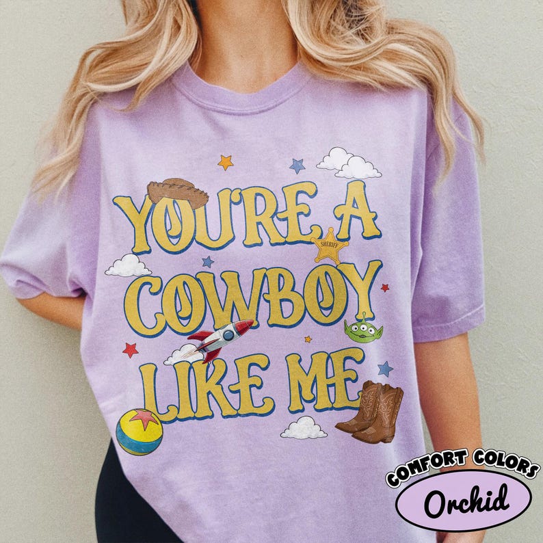 Puede incluir: Una camiseta de manga corta de color morado claro con el texto amarillo "YOU'RE A COWBOY LIKE ME" rodeado de gr&aacute;ficos que incluyen un sombrero de vaquero marr&oacute;n, una insignia de sheriff, un cohete, un alien&iacute;gena verde, una pelota y nubes.