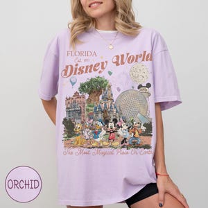 K&ouml;nnte beinhalten: Ein helllila T-Shirt mit einem Disney World-Motiv. Das Design enth&auml;lt den Text "Florida Disney World Est. 1971" sowie verschiedene Disney-Figuren und Park-Wahrzeichen. Das Shirt hat auch den Text "The Most Magical Place On Earth".
