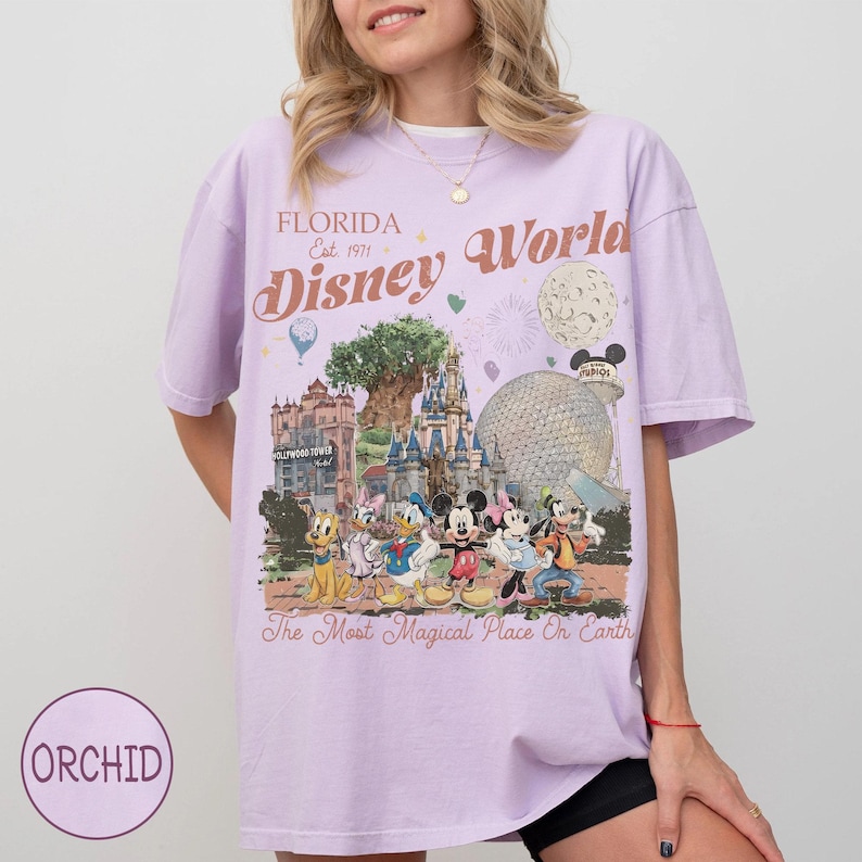 K&ouml;nnte beinhalten: Ein helllila T-Shirt mit einem Disney World-Motiv. Das Design enth&auml;lt den Text "Florida Disney World Est. 1971" sowie verschiedene Disney-Figuren und Park-Wahrzeichen. Das Shirt hat auch den Text "The Most Magical Place On Earth".
