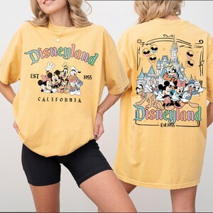 Może przedstawiać: Musztardowa koszulka z grafiką Disneyland. Prz&oacute;d koszulki przedstawia Myszka Miki, Minnie, Goofy'ego i Kaczora Donalda. Tył koszulki przedstawia zamek Disneyland i więcej postaci Disneya. Widoczny jest r&oacute;wnież tekst "Disneyland Est. 1955 California".