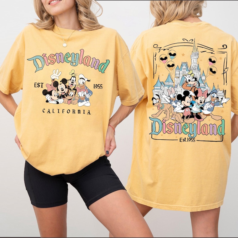 Może przedstawiać: Musztardowa koszulka z grafiką Disneyland. Prz&oacute;d koszulki przedstawia Myszka Miki, Minnie, Goofy'ego i Kaczora Donalda. Tył koszulki przedstawia zamek Disneyland i więcej postaci Disneya. Widoczny jest r&oacute;wnież tekst "Disneyland Est. 1955 California".