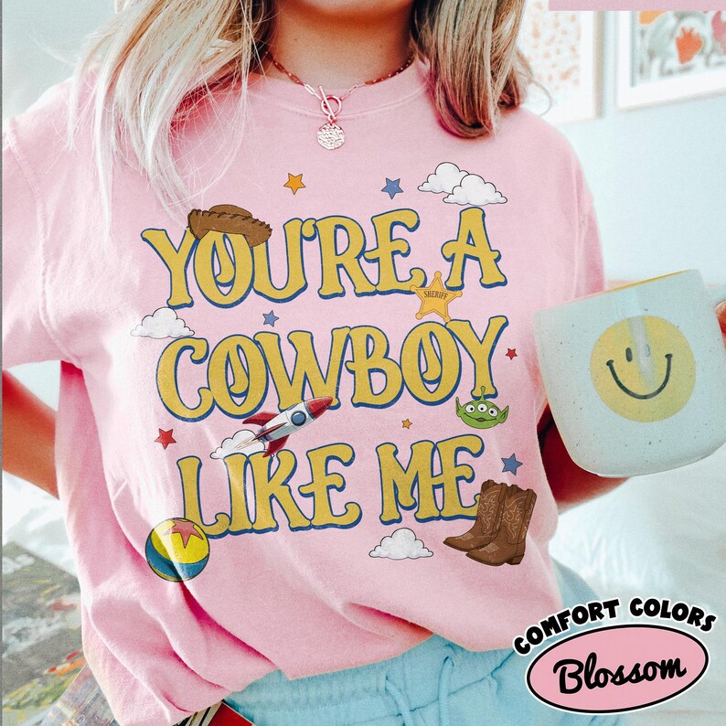 Puede incluir: Una camiseta rosa claro con el texto amarillo "YOU'RE A COWBOY LIKE ME" rodeado de gr&aacute;ficos de un sombrero de vaquero marr&oacute;n, un cohete, un alien&iacute;gena verde, una pelota colorida, una estrella de sheriff y nubes blancas, junto a una taza.