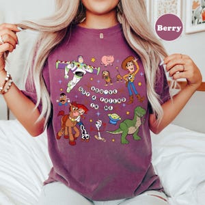 May include: Berry-coloured t-shirt with a graphic of characters from the animated film Toy Story. The design includes Buzz Lightyear, Woody, Jessie, and other characters, along with the phrase "You've Got a Friend in Me."