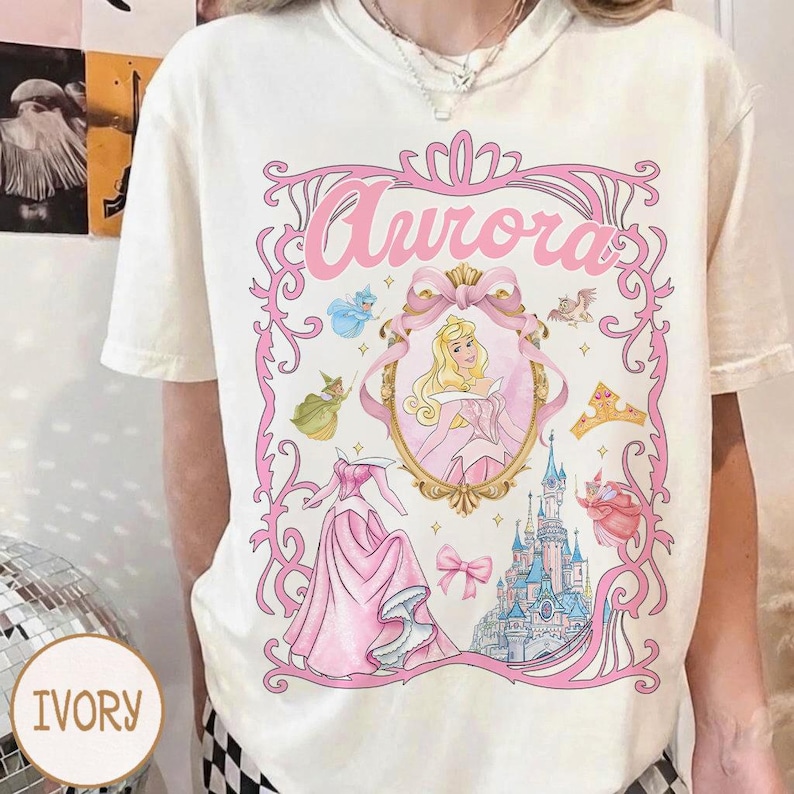 May include: Ivory t-shirt with a pink graphic of Princess Aurora. The design includes the word "Aurora", a portrait of the princess, her dress, a castle, and other fairytale elements. The shirt is a casual wear item.