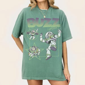 May include: Green t-shirt featuring Buzz Lightyear from Toy Story. The design includes the word "BUZZ" in gradient purple and green, along with illustrations of the character in various poses. The shirt has short sleeves and a relaxed fit.