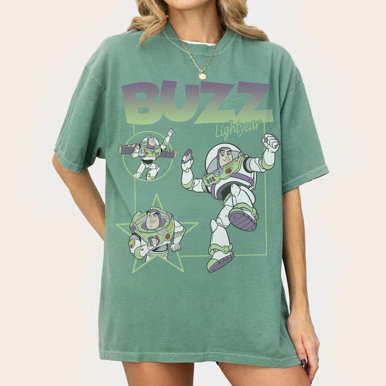 May include: Green t-shirt featuring Buzz Lightyear from Toy Story. The design includes the word "BUZZ" in gradient purple and green, along with illustrations of the character in various poses. The shirt has short sleeves and a relaxed fit.