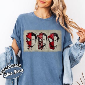 May include: Blue Comfort Colors t-shirt featuring a graphic of three Spider-Man faces. The design shows the character's mask and face in red and black, with a watercolor effect. The text "Comfort Colors Blue Jean" is on a sticker on the shirt.