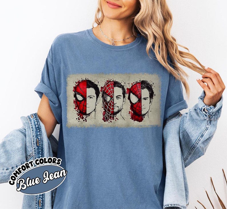 May include: Blue Comfort Colors t-shirt featuring a graphic of three Spider-Man faces. The design shows the character's mask and face in red and black, with a watercolor effect. The text "Comfort Colors Blue Jean" is on a sticker on the shirt.