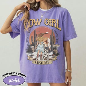 May include: A violet short-sleeve t-shirt featuring a graphic print of two figures in cowboy hats standing in a desert landscape with cacti. The text reads COW GIRL and I KNEW YOU I KNEW YOU LIKE ME. The shirt has a crew neckline and a relaxed fit.