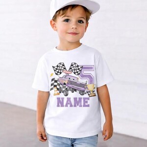 Pode incluir: Camiseta branca com um design de carro de corrida roxo, n&uacute;mero 5, bandeiras quadriculadas e um trof&eacute;u dourado. A palavra "NAME" est&aacute; em letras roxas. A camiseta &eacute; usada por uma crian&ccedil;a, ideal para um anivers&aacute;rio.