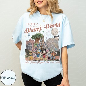K&ouml;nnte beinhalten: Hellblaues T-Shirt mit einer farbenfrohen Grafik von Disney World-Sehensw&uuml;rdigkeiten und -Charakteren. Das Design enth&auml;lt den Text "Florida Disney World" und "The Most Magical Place On Earth". Das Shirt hat kurze &Auml;rmel und einen Rundhalsausschnitt.